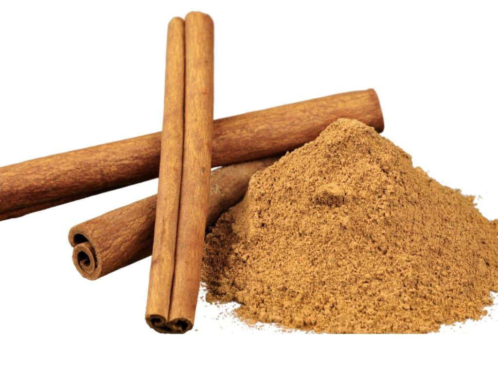 Three whole cinnamon sticks and a pile of ground cinnamon are displayed against a white background. The cinnamon sticks are slightly curved and brown, and the ground cinnamon forms a fine, light-brown powder heap—perfect for enriching your favorite autumn recipes.